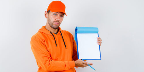 Portrait of young delivery man holding clipboard, pencil in hoodie, cap and looking confident front view