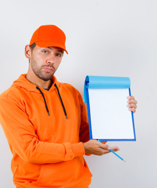 Portrait of young delivery man holding clipboard, pencil in hoodie, cap and looking confident front view